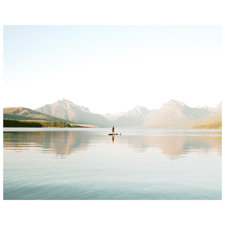 A person stands on a paddleboard with a dog on a calm lake surrounded by mountains and forest.