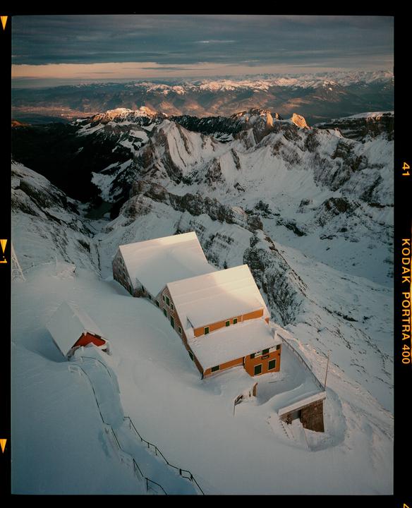 Three buildings are nestled in a snow-covered mountain landscape under a dramatic sky.