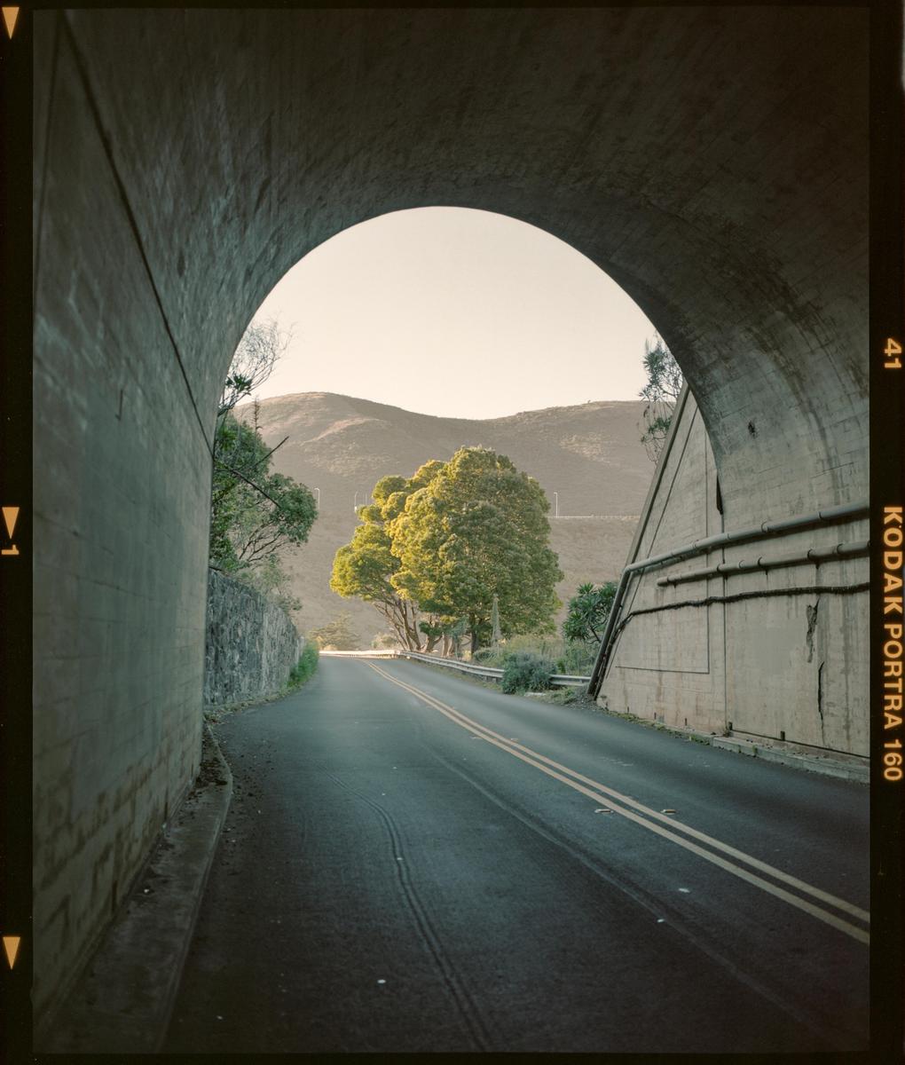 A road emerges from a concrete tunnel towards sunlit trees and mountains.