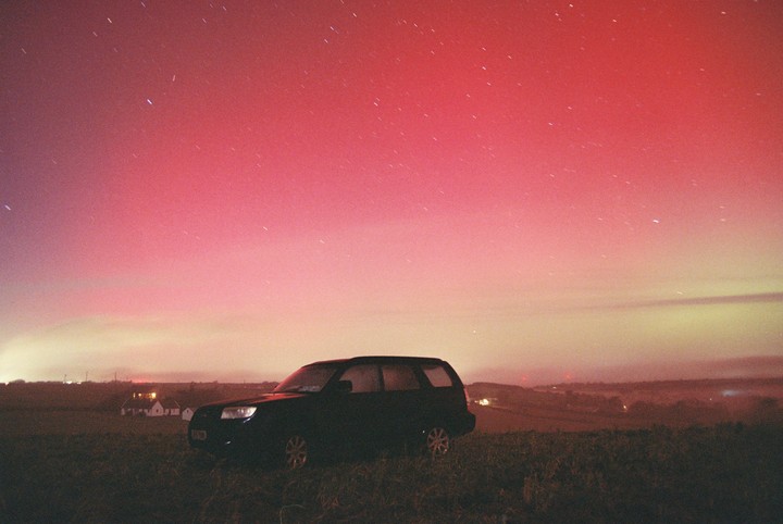 A car is parked in a field under a night sky with a red aurora borealis.