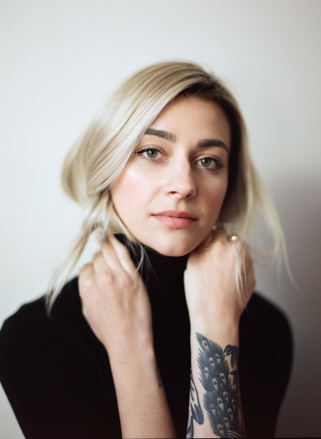 A woman with blonde hair and a black turtleneck poses with her hands near her neck, revealing a tattoo on her forearm.