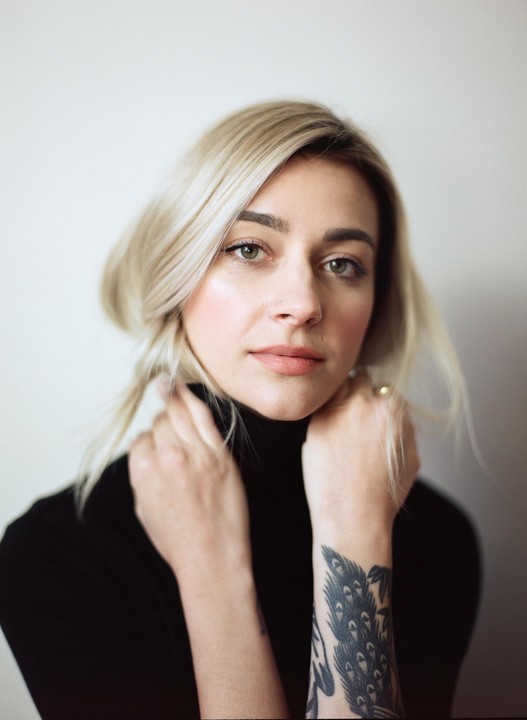A woman with blonde hair and a black turtleneck poses with her hands near her neck, revealing a tattoo on her forearm.