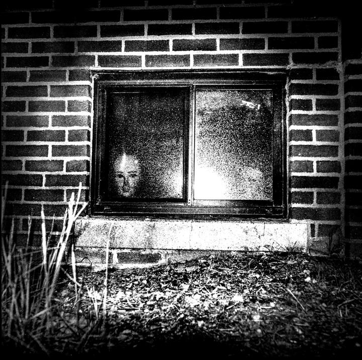 A face is visible through a grimy basement window in a brick wall.