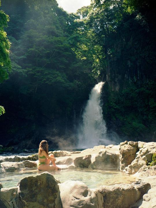 A woman sits in a hot spring with a waterfall and lush forest in the background.