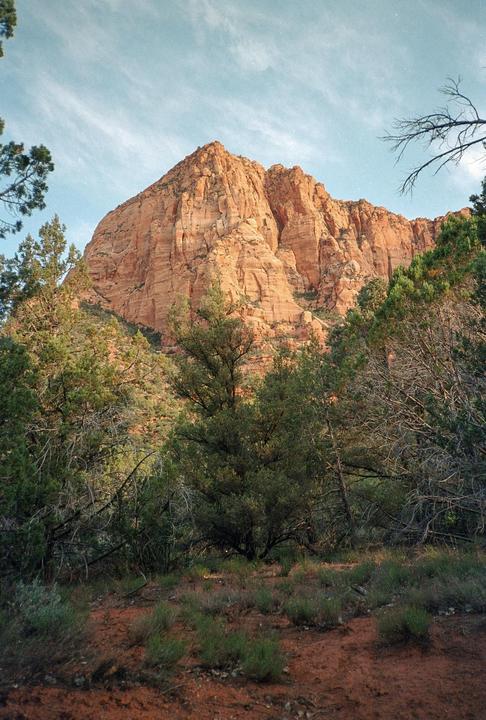A large red rock mountain rises above green trees and shrubs under a blue sky with wispy clouds.
