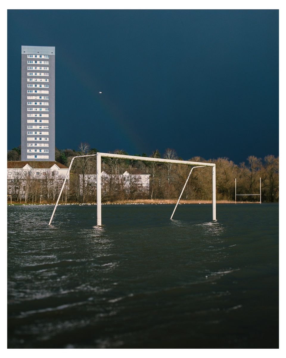A flooded sports field with a soccer goal and rugby goal is visible in the foreground, with a tall building and trees in the background under a dark sky.
