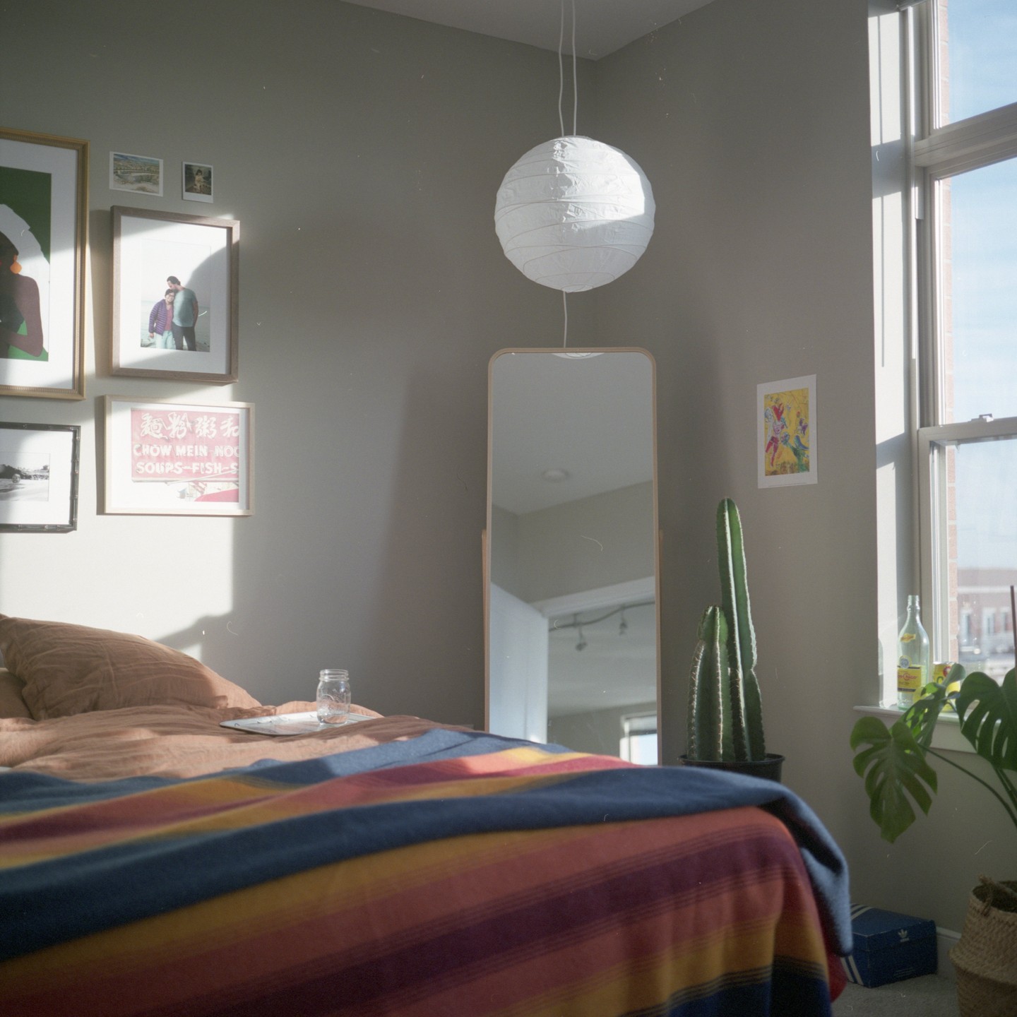 Sunlight streams into a bedroom with a striped blanket on the bed, a paper lantern, and a tall mirror.