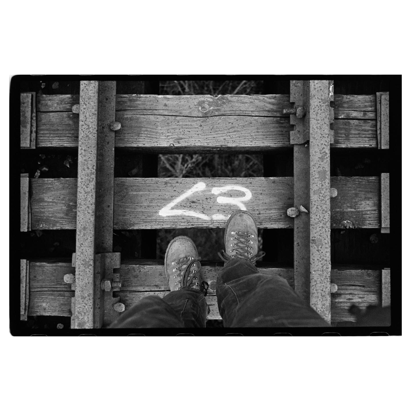 A person's feet are visible on a wooden railway bridge with a painted symbol.