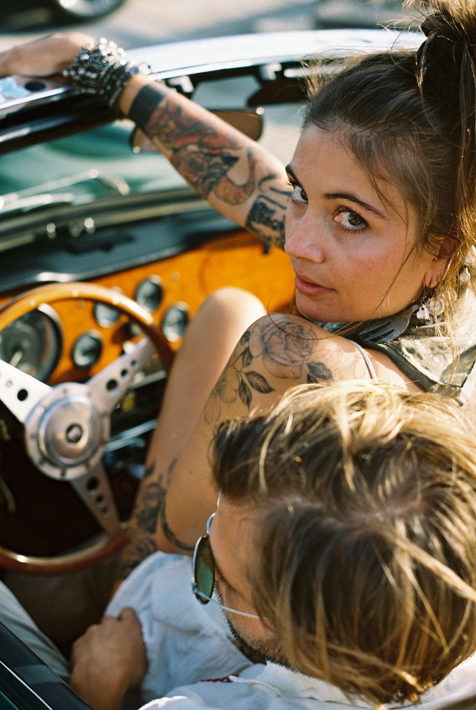 A woman with tattoos on her arms looks back at the viewer from the passenger seat of a convertible car, with a man in the driver's seat.