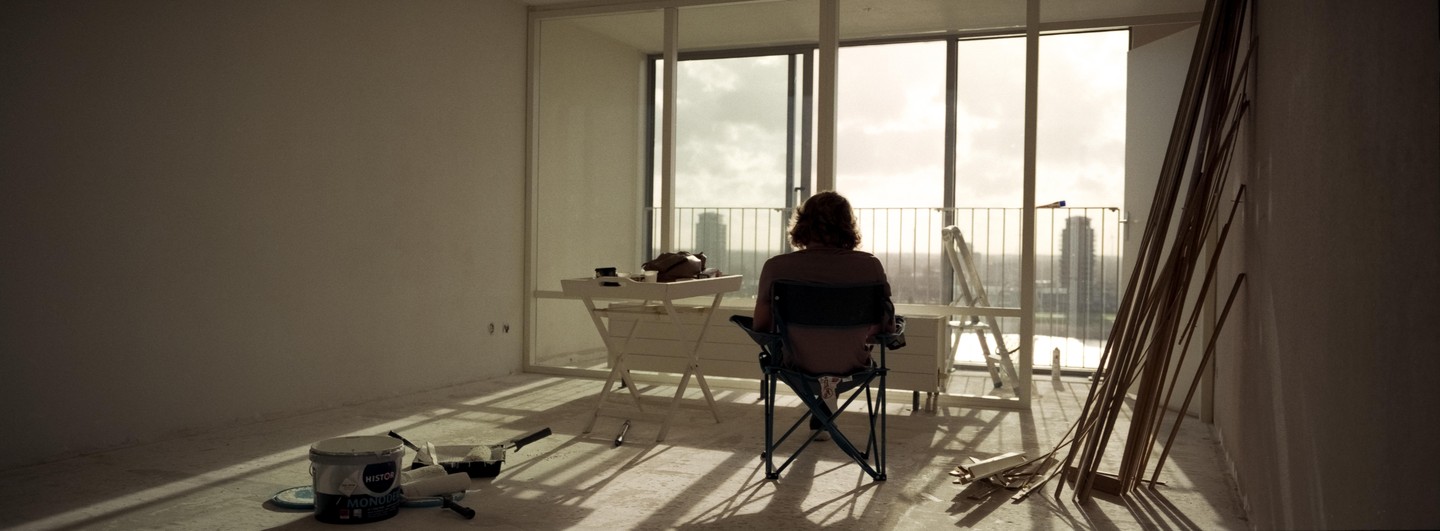 A person sits in a folding chair looking out a large window at a city skyline from an empty room with painting supplies and wood trim.