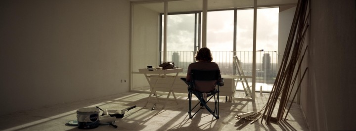 A person sits in a folding chair looking out a large window at a city skyline from an empty room with painting supplies and wood trim.