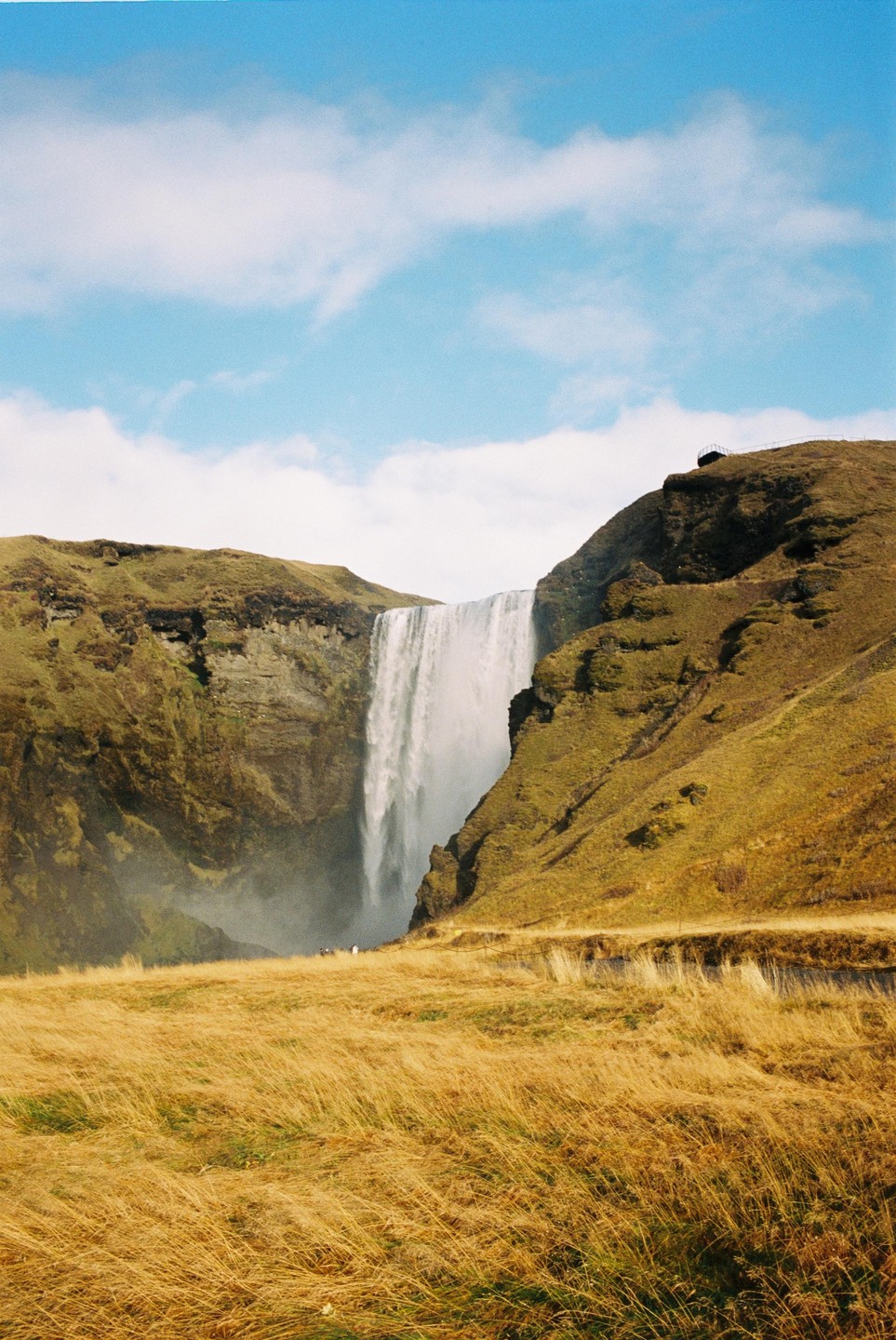 A large waterfall cascades down a cliff into a misty pool, surrounded by grassy hills under a blue sky with clouds.