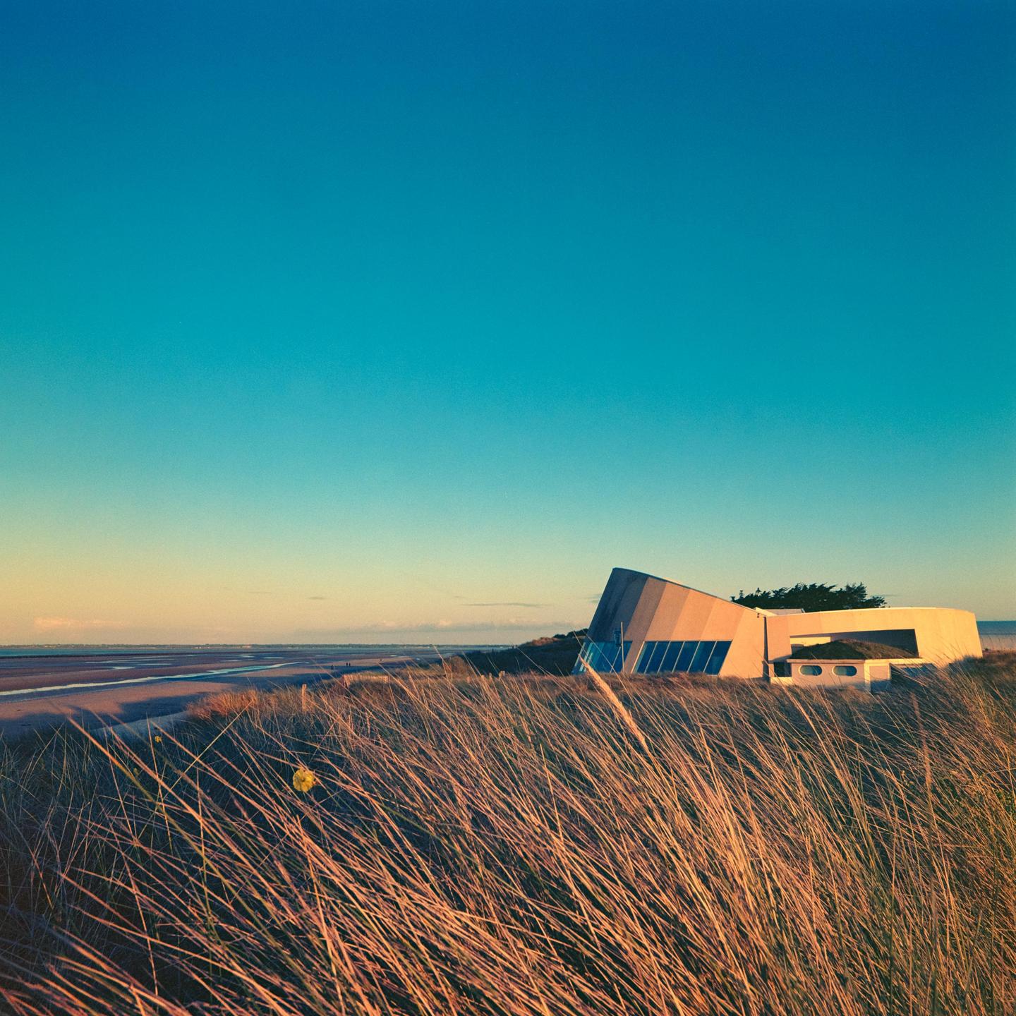 A modern building with a sloped roof stands on a grassy dune overlooking a beach at sunset.