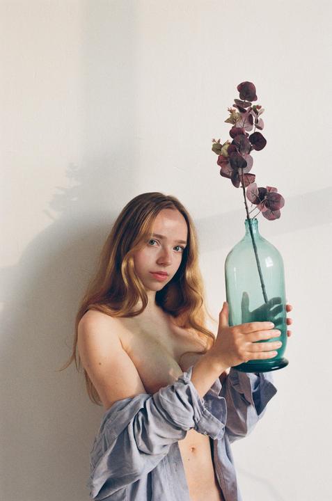 A woman with long hair holds a large glass vase with a plant inside.
