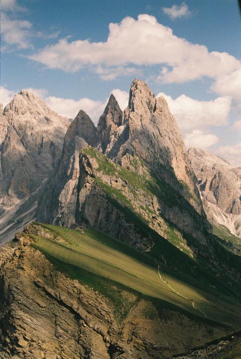 A mountain range with jagged peaks and green slopes is visible under a blue sky with white clouds.