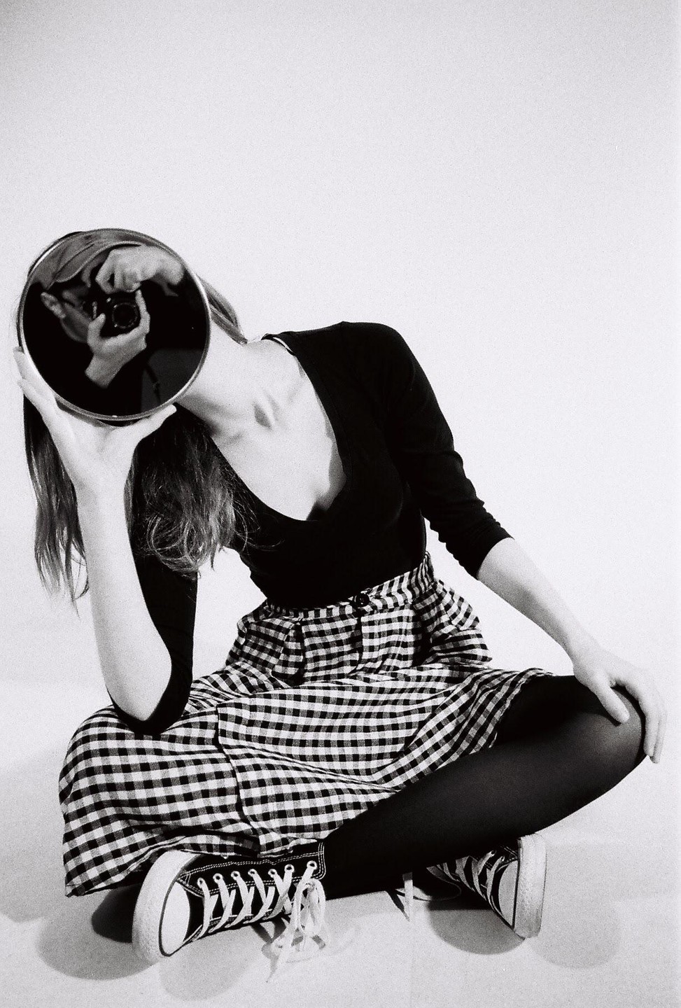A woman sits on the floor, holding a mirror that reflects her face and camera.