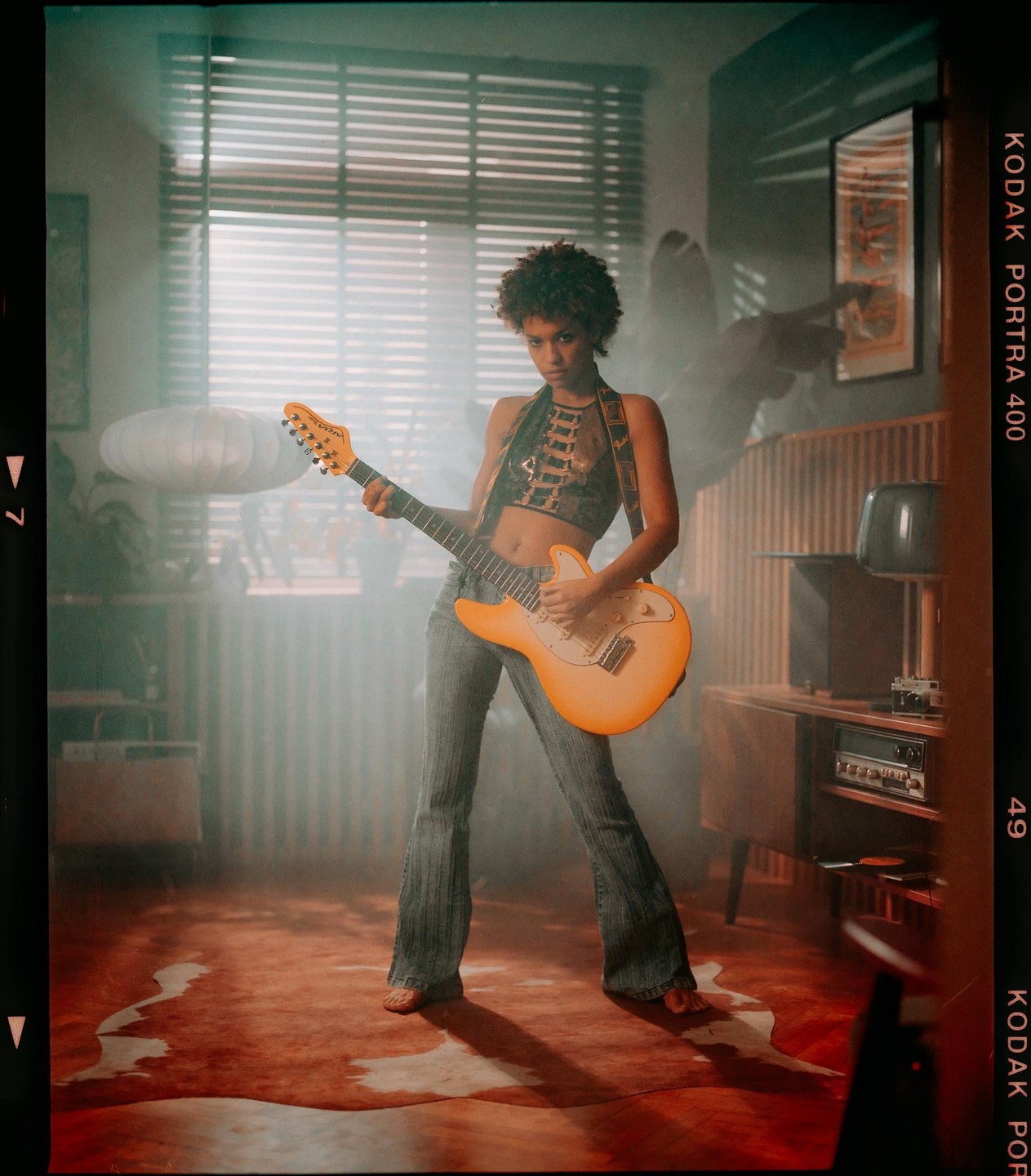 A woman with an orange electric guitar stands barefoot in a room with a window and a record player.