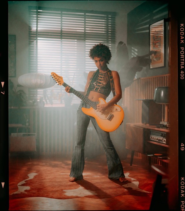 A woman with an orange electric guitar stands barefoot in a room with a window and a record player.