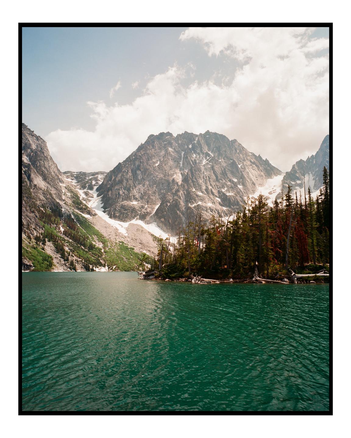 A turquoise lake is surrounded by a forest and snow-capped mountains under a cloudy sky.