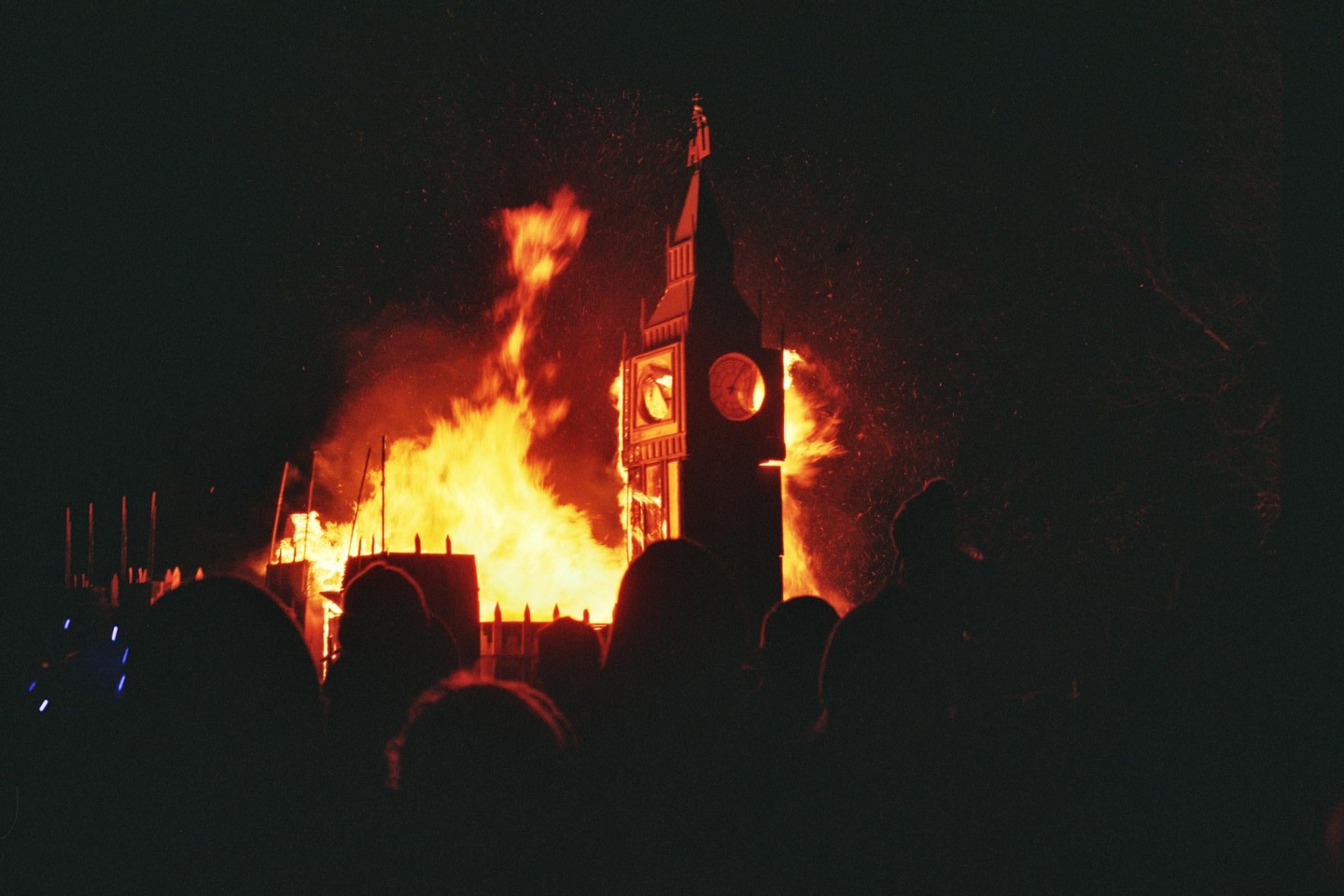 A crowd of people watches a large fire engulfing a clock tower at night.