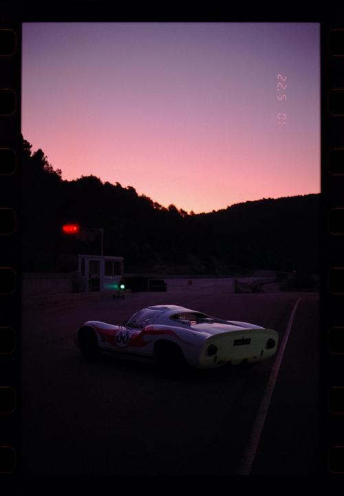 A white race car with red and orange stripes is parked on a track at dawn, with a vibrant pink and purple sky overhead.