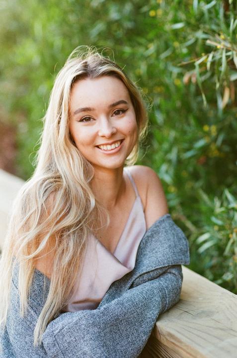 A woman with long blonde hair smiles at the viewer while leaning on a wooden railing.