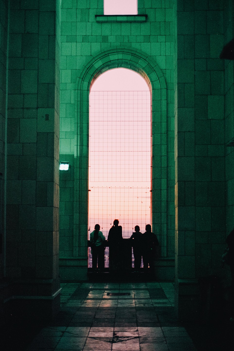 Four people are silhouetted against a large arched window with a grid pattern, looking out at a pink sky.