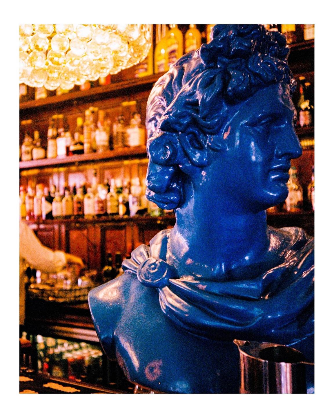 A blue bust of a man is on a bar counter with bottles of alcohol in the background.