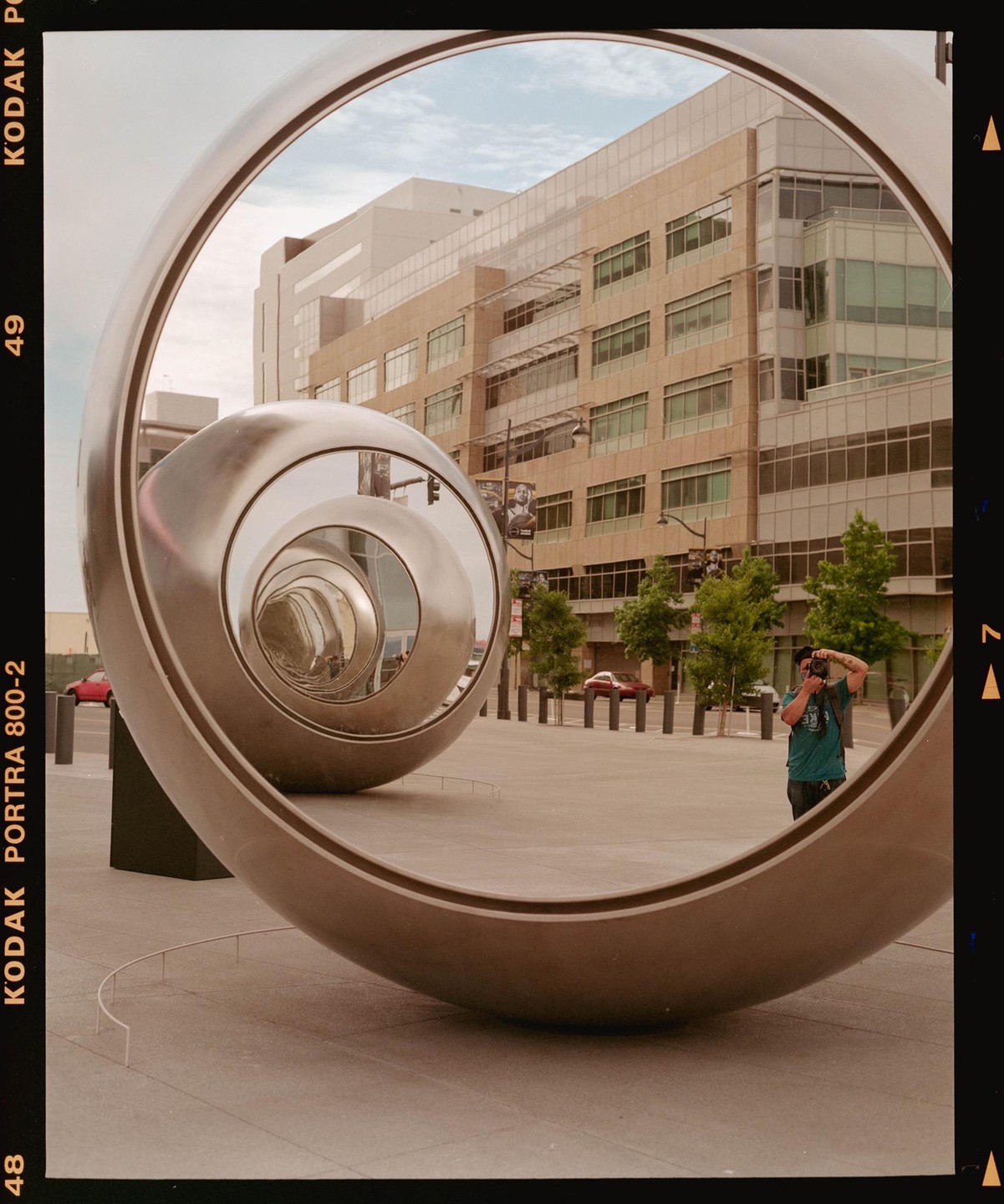 A man takes a self-portrait reflected in a large, spiraling metal sculpture in a city square, with a building and trees in the background.