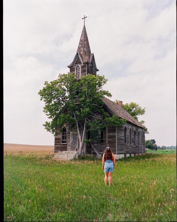 A woman stands in a field of tall grass, looking at an old wooden church with a tall steeple.
