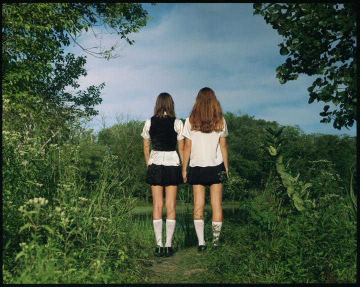 Two girls stand with their backs to the viewer, looking out at a lagoon surrounded by lush green foliage.