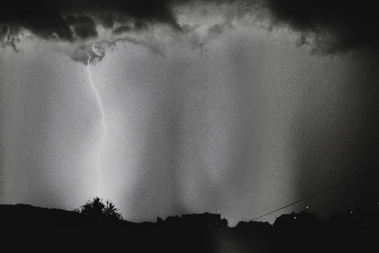 A bolt of lightning strikes from dark clouds above a silhouetted landscape with trees and buildings.