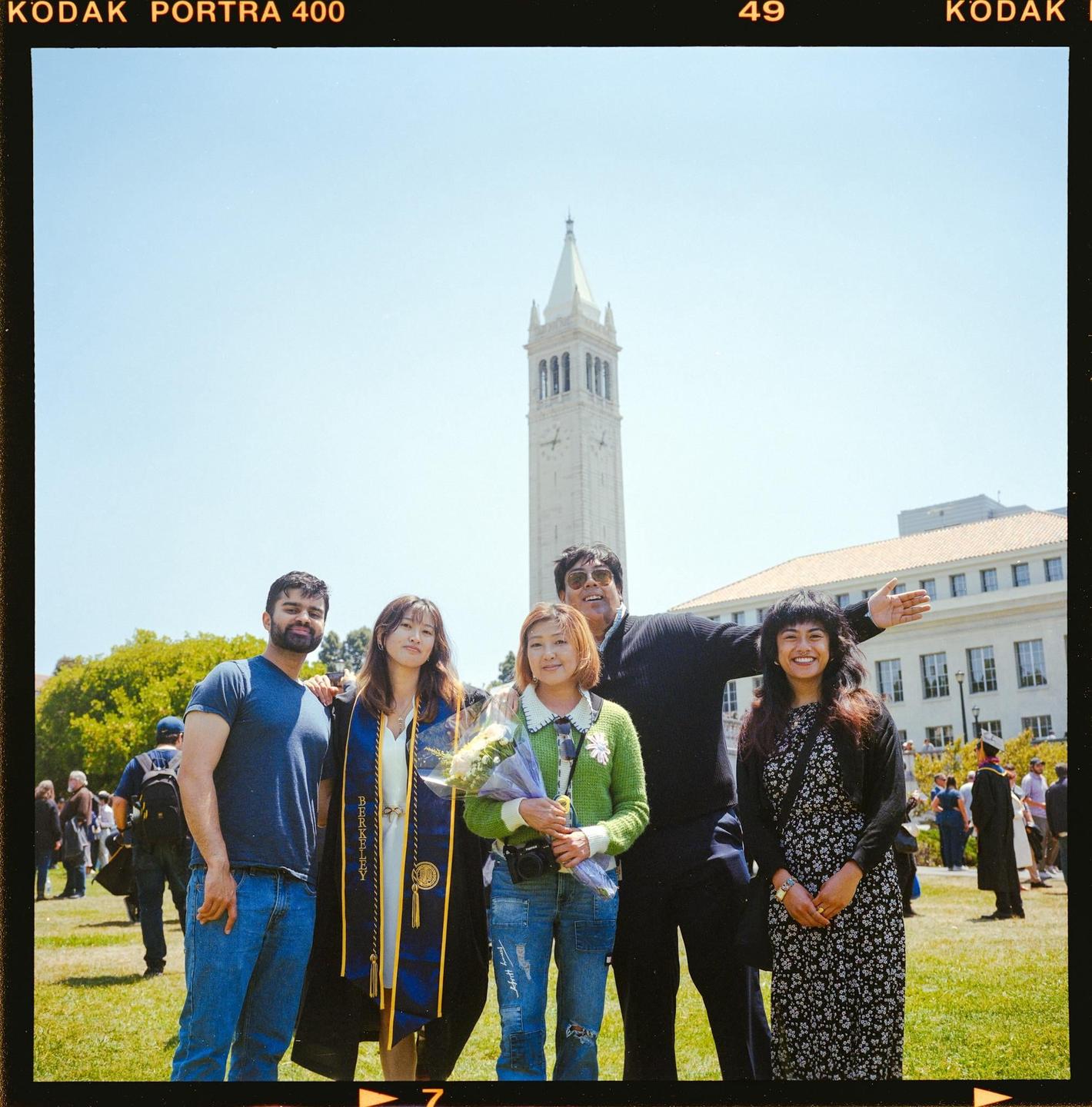 A group of five people stand on a grassy lawn in front of a tall clock tower and a building.