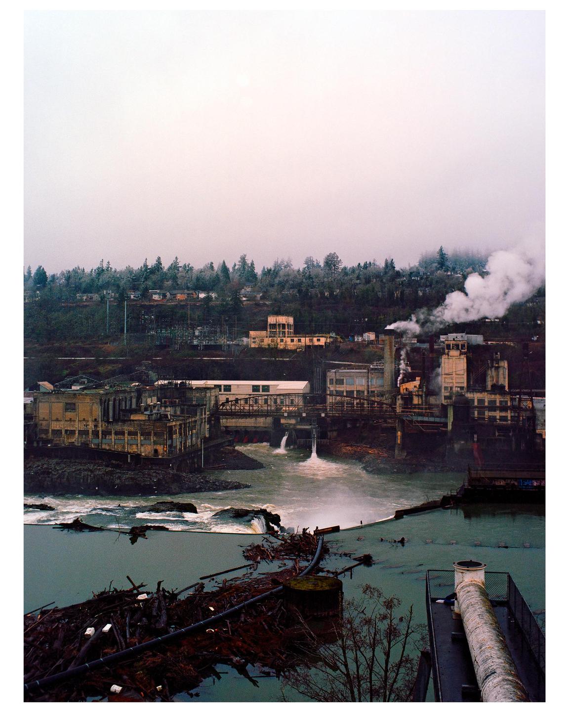 A river flows past industrial buildings and a forested hill under a hazy sky.