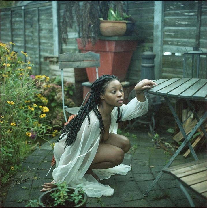 A woman in a sheer white top crouches next to a table in a garden.