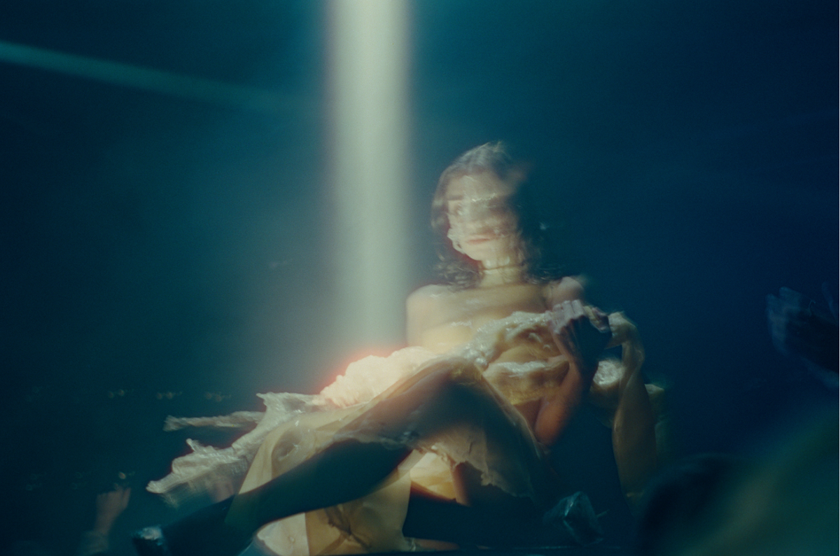 A woman with blurred features is illuminated by a beam of light from above while sitting in water.
