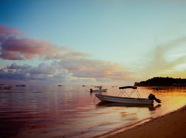 A small boat is moored on a sandy beach with a calm ocean and a colorful sunset sky.