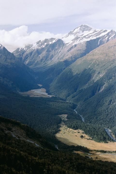 A valley with a river and forest is nestled between snow-capped mountains under a cloudy sky.