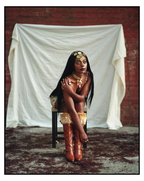 A woman with long dark hair and a coin headpiece sits on a stool in front of a white sheet and brick wall.
