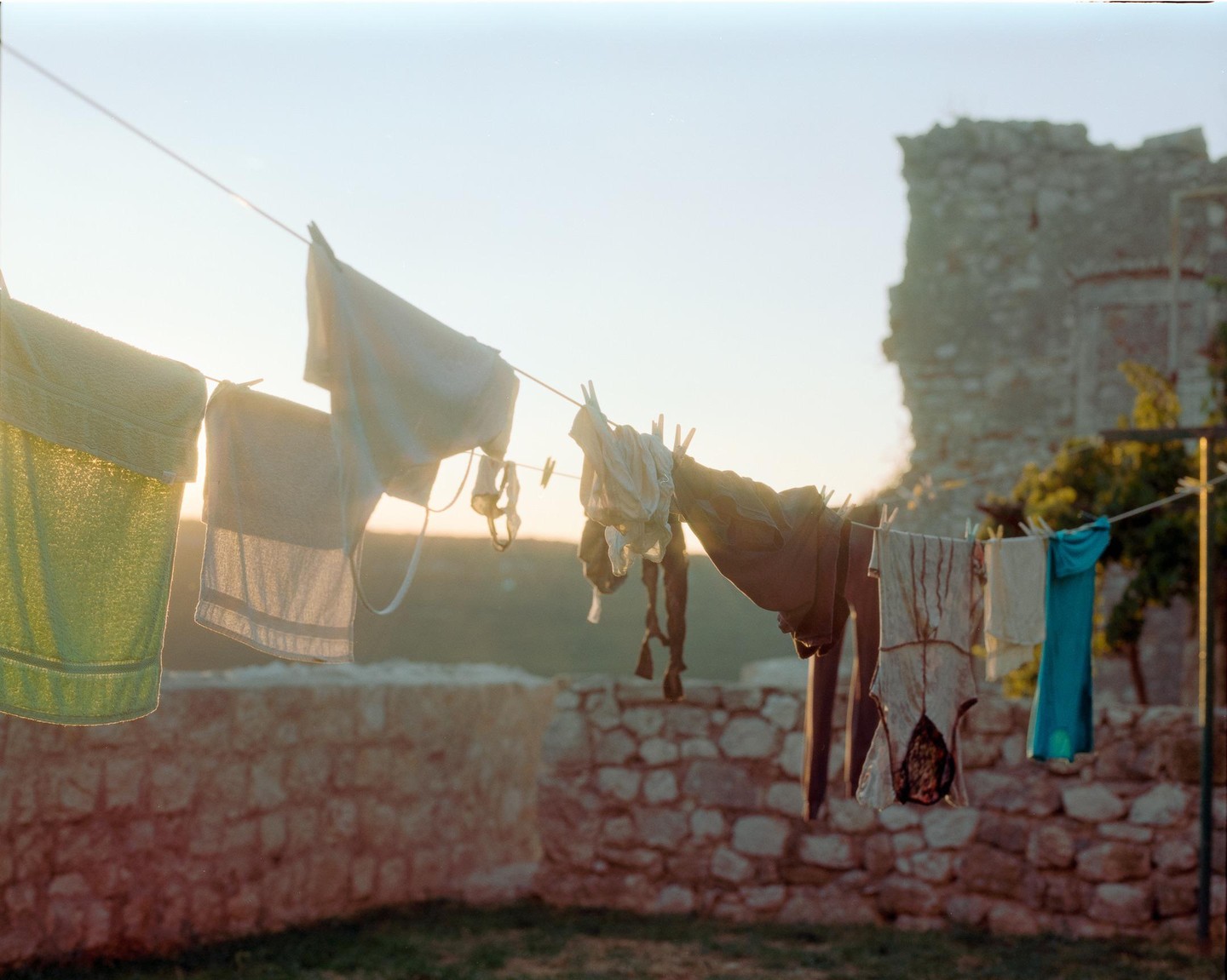 Laundry hangs on a clothesline in front of a stone wall and a ruined building at sunset.