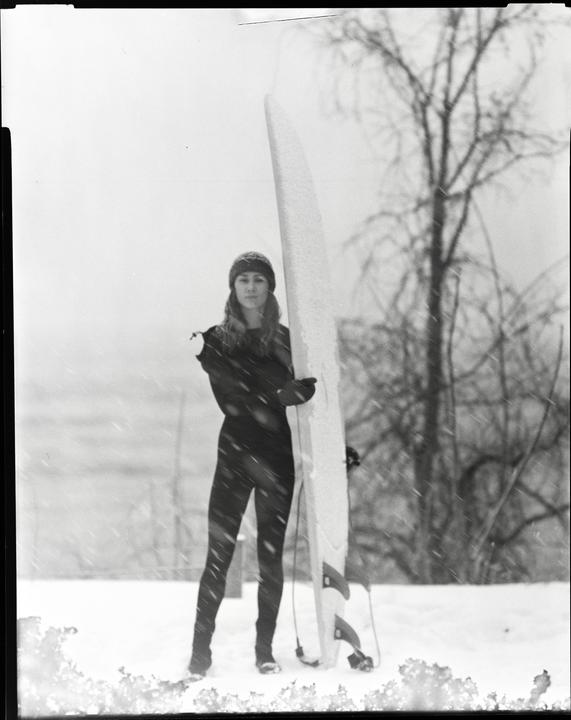 A woman in a wetsuit and beanie stands in the snow next to a surfboard.