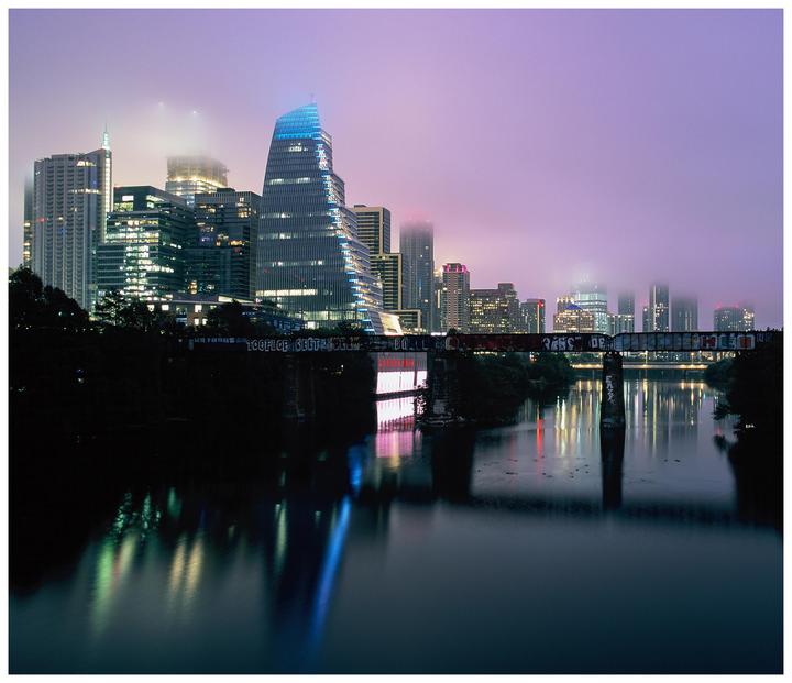 A city skyline with illuminated buildings is reflected in a river under a hazy purple sky at night.