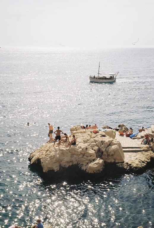 People are swimming and sunbathing on rocks in the Adriatic Sea, with a boat in the distance.
