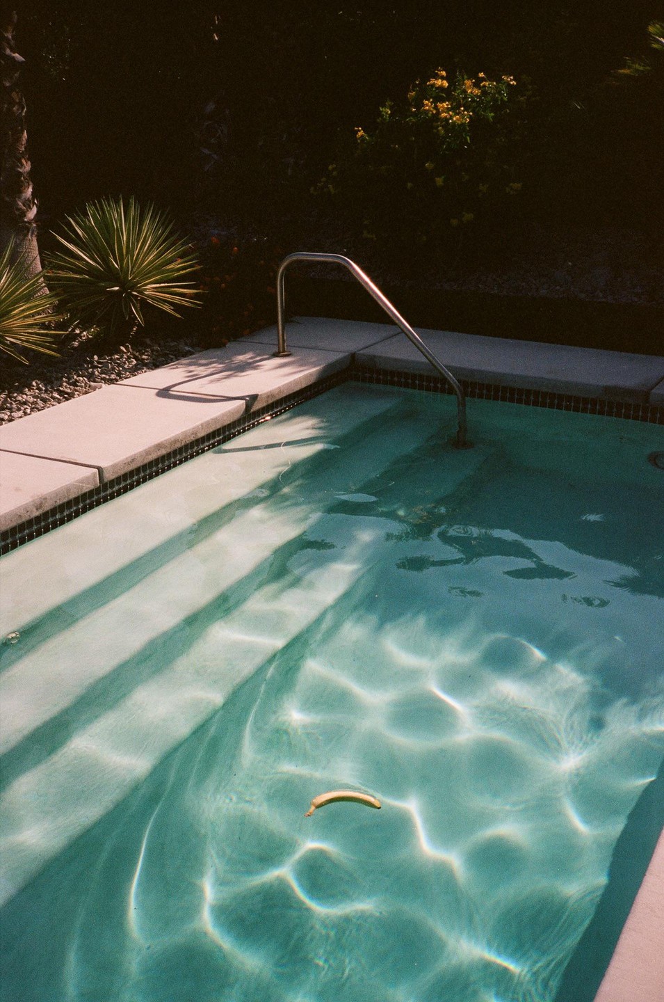 A banana floats in a swimming pool with steps and a handrail, surrounded by plants.