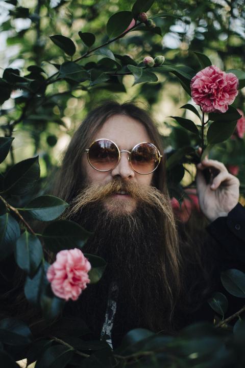 A man with a long beard and round sunglasses peeks through green foliage with pink flowers.