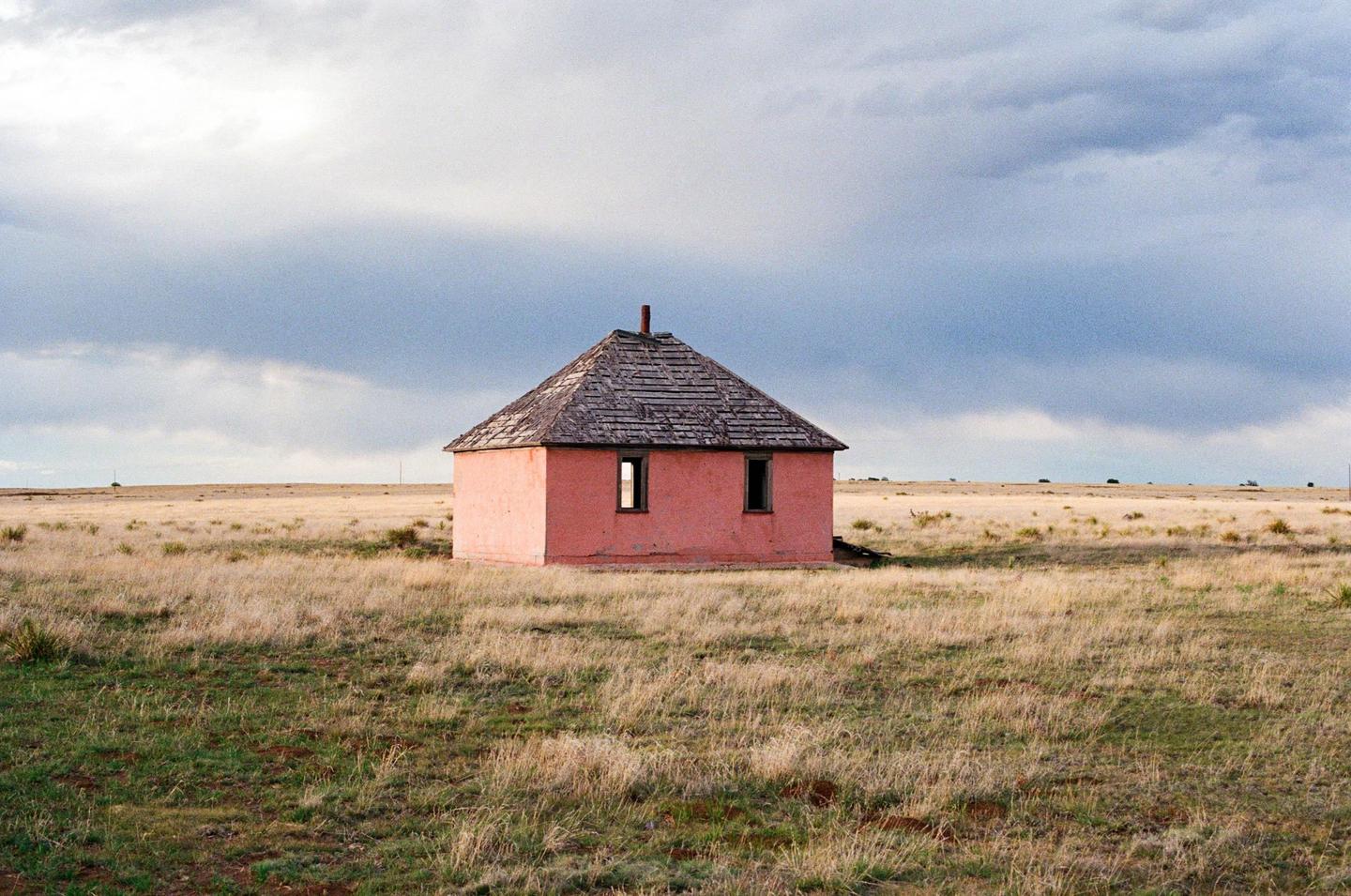A small pink house with a dark roof stands in a field of dry grass under a cloudy sky.
