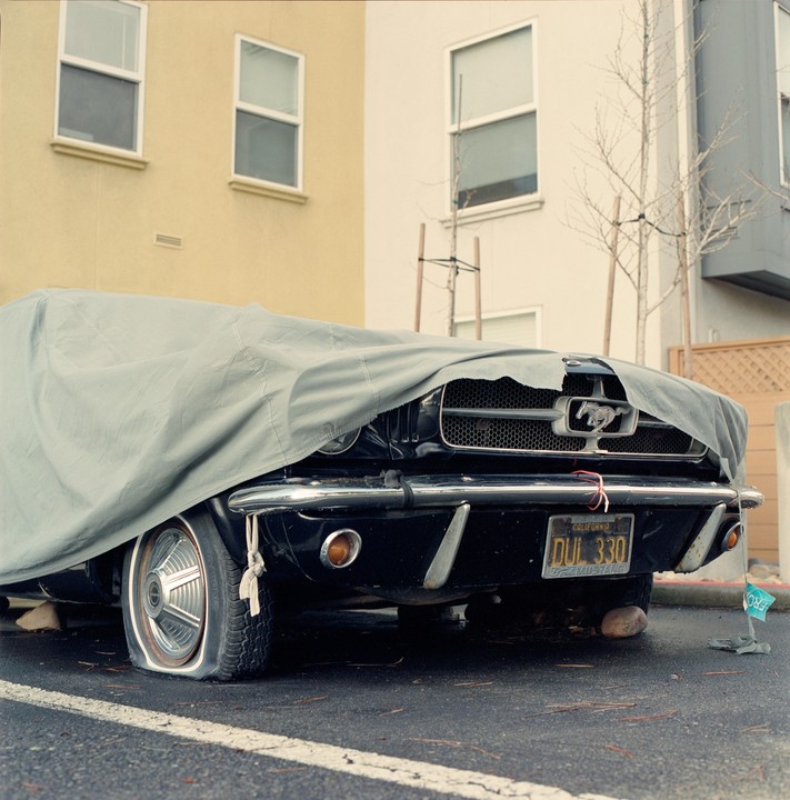 A black Mustang with a flat tire is partially covered by a grey tarp, parked on asphalt in front of buildings.