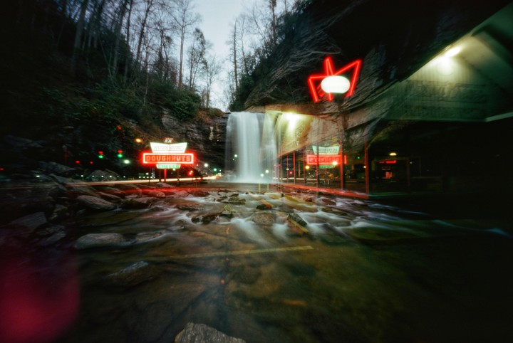 A waterfall cascades next to a road with neon signs for doughnuts and coffee at night.