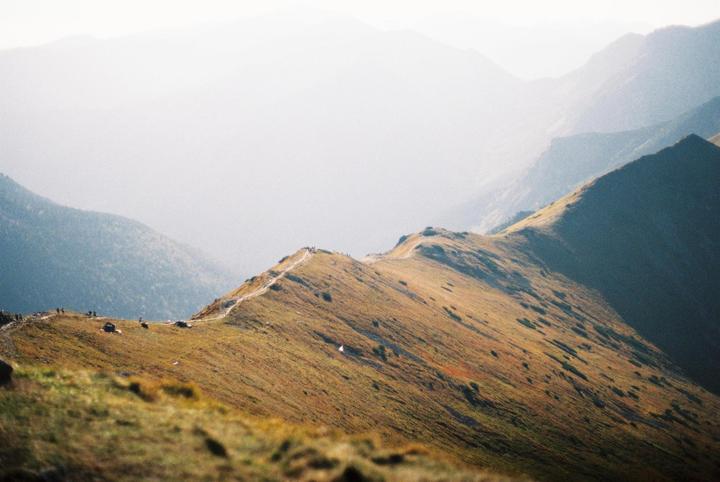 A winding path leads across a mountain ridge with other mountains in the hazy background.