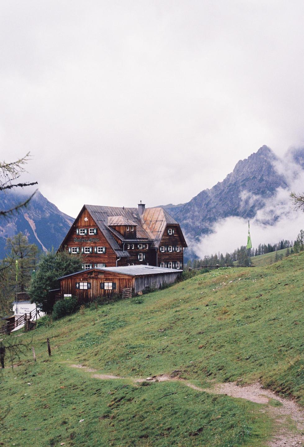 A large wooden building with green shutters sits on a grassy hill with mountains and clouds in the background.
